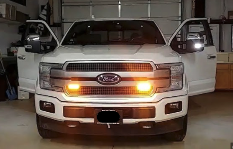 A white Ford truck with headlights illuminated, parked inside a spacious garage.